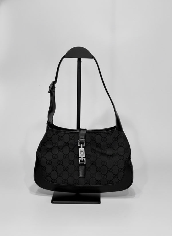 Gucci Jackie black canvas shoulder bag featuring the iconic GG monogram pattern, leather trim, and silver-tone piston clasp, photographed on a display stand.